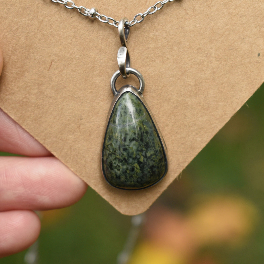 Teardrop-shaped green stone pendant on a silver chain held by a hand against a brown background.