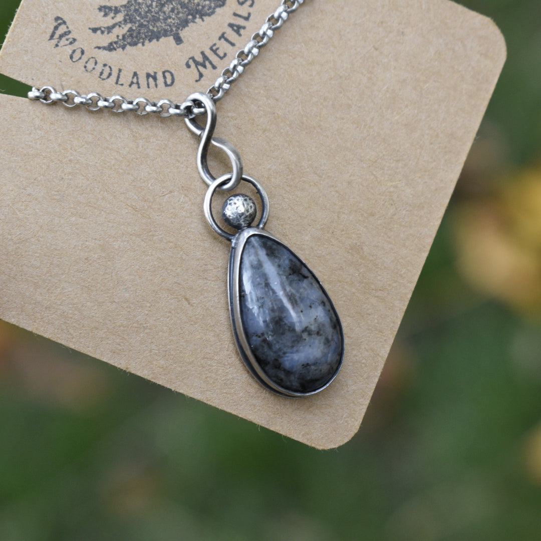 Necklace with a teardrop-shaped pendant on a Woodland Metalsmith card against a blurred natural background.