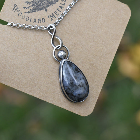 Necklace with a teardrop-shaped pendant on a Woodland Metalsmith card against a blurred natural background.