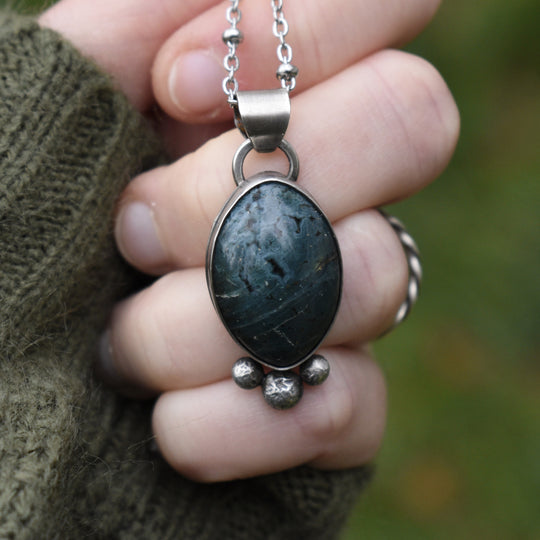 Necklace with a dark stone pendant held between fingers against a blurred green background