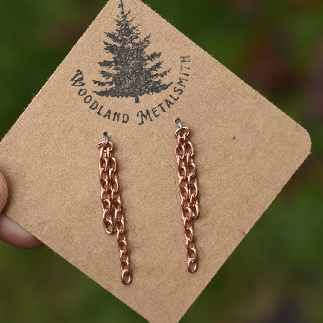 Rose gold chain earrings on a 'Woodland Metalsmith' branded card with a blurred green background.