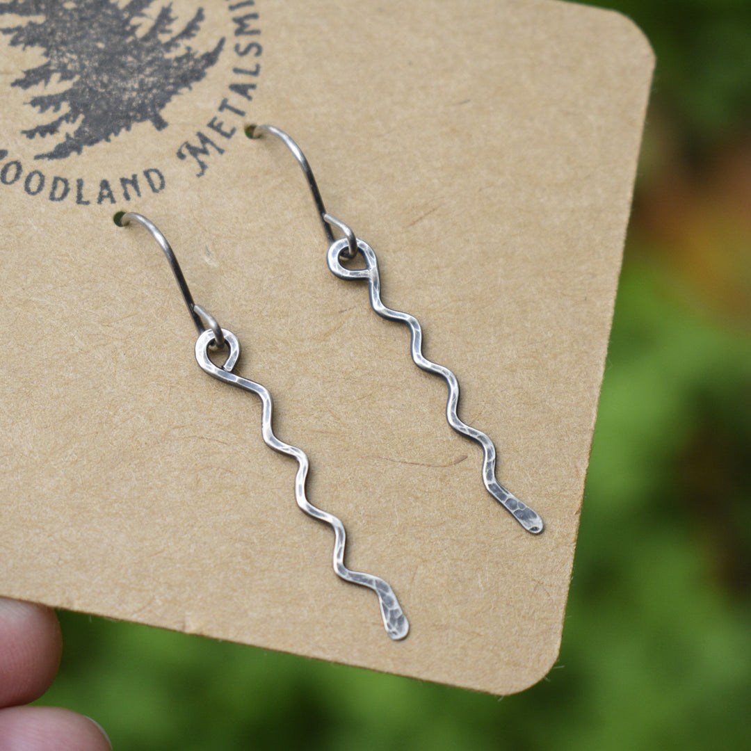 Silver wavy earrings on a 'Woodland Metalsmith' branded card with a blurred natural background.