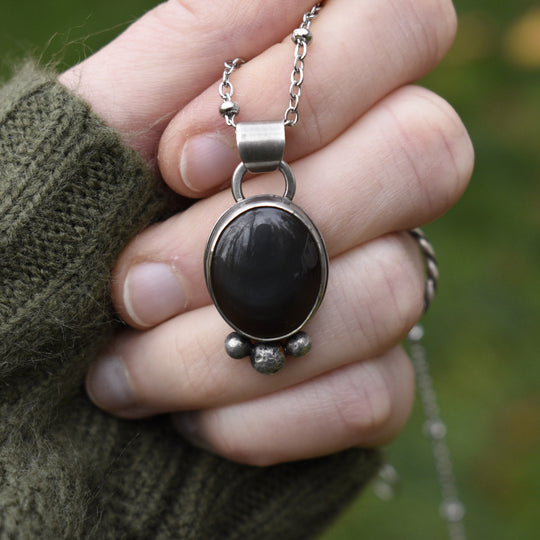 Hand holding a silver necklace with a black pendant against a blurred green background