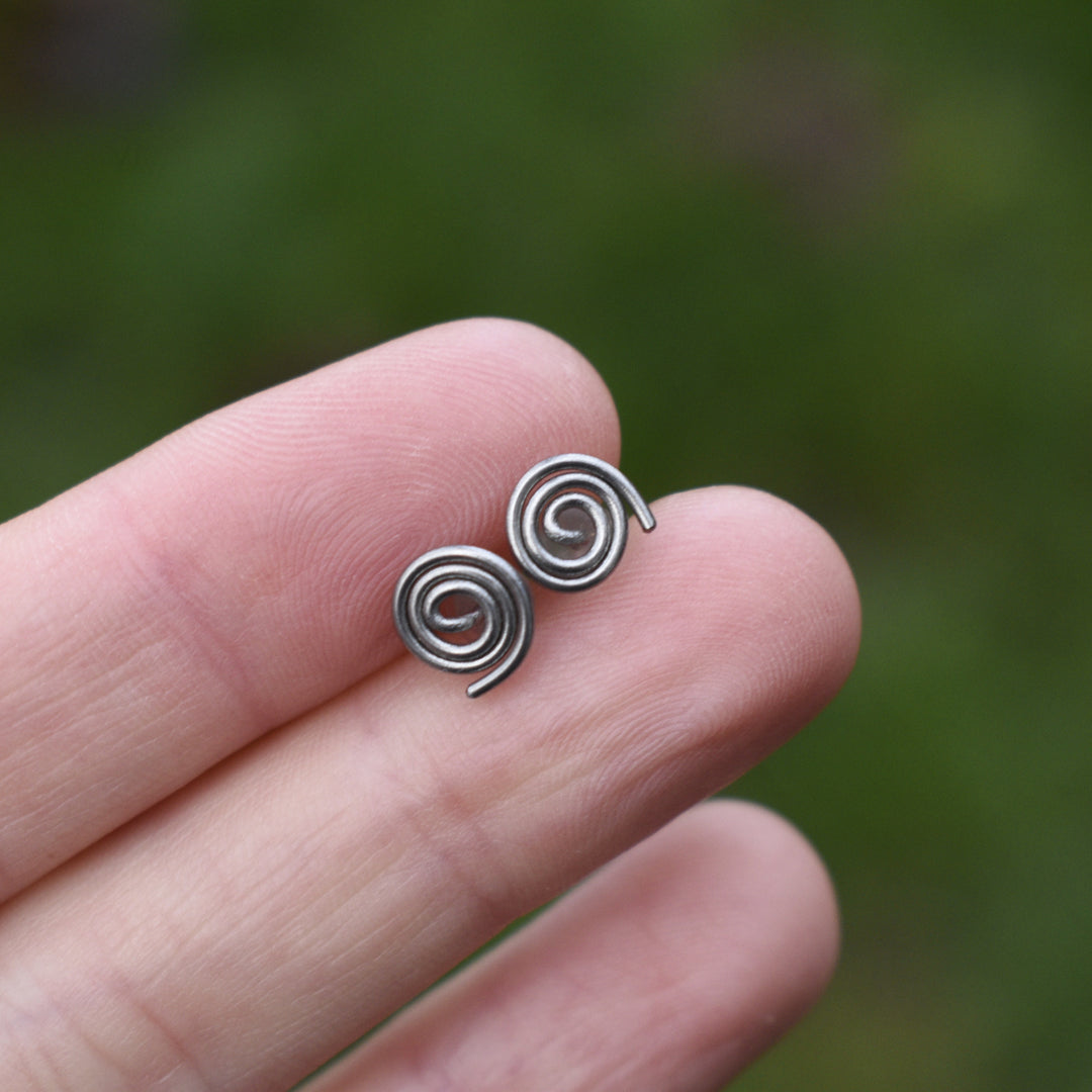 Silver spiral earrings held between two fingers against a blurred green background